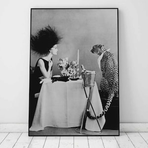 May include: A black and white photograph of a woman wearing a large black feathered hat sitting at a table with a cheetah standing behind her. The woman is wearing a black dress and the cheetah is wearing a white collar. There is a champagne bucket and glasses on the table.