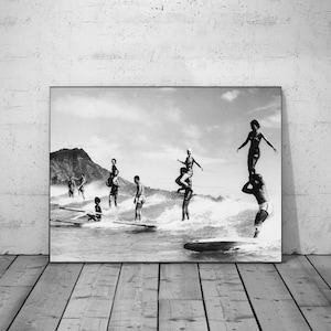 Op de afbeelding: Zwart-witfoto van surfers op golven. Verschillende surfers staan op surfplanken, sommigen met anderen op hun schouders. De afbeelding toont een surfcène met een berg op de achtergrond.