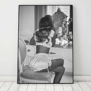 Op de afbeelding: Een zwart-witfoto van een persoon die voor een kaptafelspiegel zit, gekleed in een wit satijnen lingerie-set. De persoon kijkt naar hun reflectie in de spiegel.