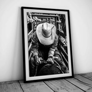 May include: Black and white photo of a cowboy riding a bull in a rodeo. The cowboy is wearing a wide-brimmed hat and a long-sleeved shirt. The bull is bucking and the cowboy is holding on tight.