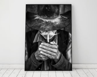 May include: Black and white framed print of a cowboy lighting a cigarette. The cowboy wears a hat and a jacket. The image is a close-up of the cowboy's hands and face, with a cigarette in his mouth.