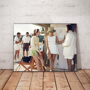 May include: A framed print depicting a vintage tennis scene. The image shows several people, some in tennis attire, gathered around a diagram. The color palette is soft, with a focus on whites, greens, and browns, evoking a sense of nostalgia.