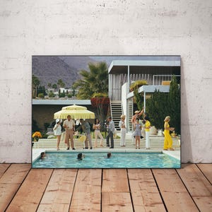 May include: A framed photographic print depicting a mid-century modern house with a swimming pool and a group of people socializing outdoors. The scene features a yellow umbrella, palm trees, and individuals dressed in colorful summer attire.