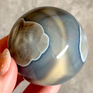 May include: A spherical gemstone, polished to a high sheen, displaying a blend of grey, white, and brown tones. The orb has cloud-like patterns. This decorative sphere is a unique home decor item.