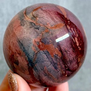 May include: A polished sphere in shades of burgundy, reddish-brown, and dark grey. The surface has a textured appearance with natural patterns and veins. The sphere is smooth and reflective, with a glossy finish, and is likely made of stone.