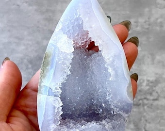 Peut inclure: Une géode d'agate bleu clair avec une grande ouverture révélant un intérieur tapissé de cristaux. La géode a la forme d'une pointe et présente un extérieur lisse et poli.