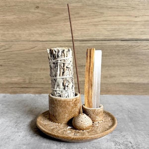 May include: A brown ceramic incense holder with a bundle of white sage, a stick of palo santo, and an incense stick. The holder has a speckled design and a small, round incense holder. The base is a round plate with small holes.