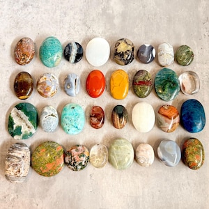May include: A collection of thirty-two polished oval gemstones arranged in four rows. The stones feature a variety of colours including green, orange, blue, white, brown, and grey, with diverse patterns such as speckles, stripes, and translucent layers.