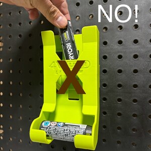 Puede incluir: Un soporte para bater&iacute;as de pl&aacute;stico verde con el texto "4VUS" y "RYOBI" impreso en &eacute;l. Una bater&iacute;a est&aacute; parcialmente insertada en el soporte. El texto "NO!" est&aacute; escrito en blanco sobre un fondo negro.