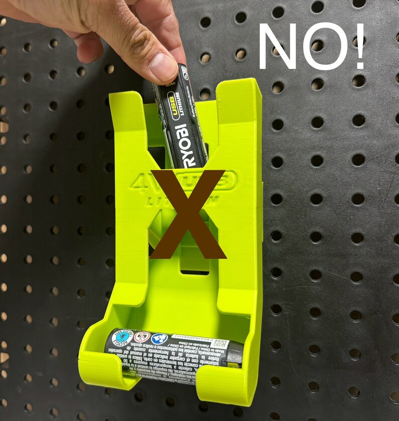 Puede incluir: Un soporte para bater&iacute;as de pl&aacute;stico verde con el texto "4VUS" y "RYOBI" impreso en &eacute;l. Una bater&iacute;a est&aacute; parcialmente insertada en el soporte. El texto "NO!" est&aacute; escrito en blanco sobre un fondo negro.