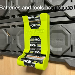 Puede incluir: Un soporte de bater&iacute;a impreso en 3D de color verde para bater&iacute;as de litio Ryobi de 4V. El soporte est&aacute; montado en una pared negra y tiene capacidad para cuatro bater&iacute;as. El texto "Batteries and tools not included" es visible en la parte superior de la imagen.