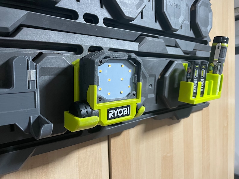 以下が含まれることがあります： 黒と緑のRyobi作業灯。白いLEDライトパネルと吊り下げ用のフックが付いています。ライトは、Ryobiバッテリー用のバッテリーホルダーが付いた黒いペグボードに取り付けられています。