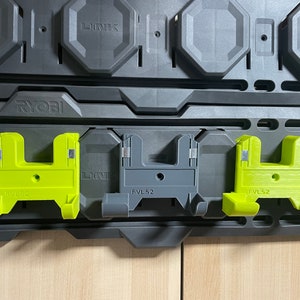 以下が含まれることがあります： Ryobi ツールシステム用のツールホルダー3個。ホルダーはプラスチック製で、緑とグレーです。ホルダーには「FVL52」と「RYOBI」のラベルが付いています。