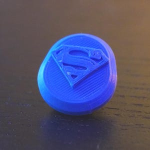 May include: A small, circular, blue plastic button featuring a raised Superman logo in the center. The surface of the button has a visible, fine, horizontal 3D-printed texture across the entire face, including the embossed letter S emblem.