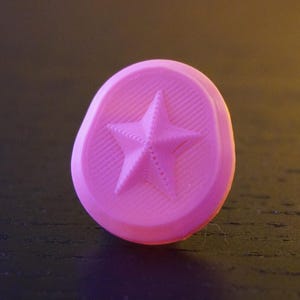 Può includere: Un bottone circolare rosa con un design a stella a cinque punte in rilievo al centro. La superficie del bottone presenta una sottile trama lineare che si irradia dalla stella e i bordi sono leggermente irregolari e arrotondati.