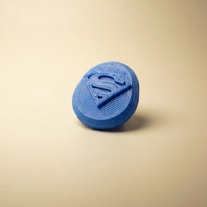 May include: A circular blue plastic object featuring a raised Superman logo in the center. The surface of the disc displays visible horizontal 3D printed layer lines, and the emblem is embossed with a diamond-shaped border and the letter S.