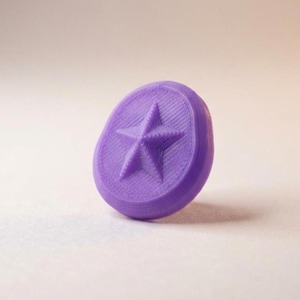 May include: A circular purple button featuring a raised five-pointed star design in the center. The surface of the button displays a textured, layered finish consistent with 3D printing, with a smooth, rounded edge surrounding the star motif.