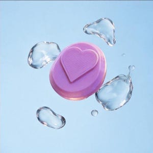 May include: A circular lavender object featuring a raised heart shape on its surface, surrounded by several clear, liquid-like droplets suspended against a solid light blue background. The object has a smooth, matte texture and a distinct heart icon.