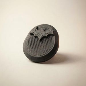 Puede incluir: Un objeto negro, circular, impreso en 3D con una superficie texturizada que presenta un emblema en relieve con forma de murciélago en la parte superior. El artículo tiene una apariencia estratificada distintiva del proceso de impresión.