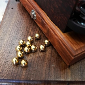 Puede incluir: Una colección de pequeñas esferas metálicas de color dorado pulido esparcidas sobre una superficie de madera marrón oscuro junto a una base de madera con un botón circular de tono plateado. La base de madera presenta una veta natural rica.