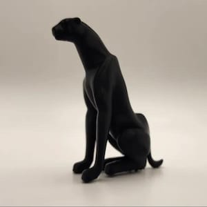 May include: A black panther statue in a seated position, with its head turned slightly to the left. The sculpture has a smooth, matte finish and is set against a plain background. The panther's pose suggests alertness.