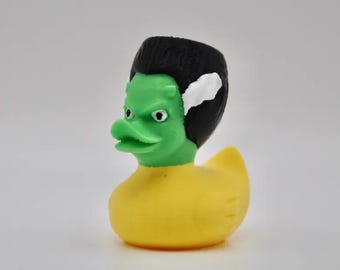 Könnte beinhalten: Eine neuartige Gummiente mit Frankenstein-Monster-Thema. Die Ente hat ein grünes Gesicht, schwarzes Haar und einen gelben Körper. Das Gesicht der Ente hat einen strengen Ausdruck.