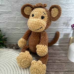 Könnte beinhalten: Ein handgefertigtes, plüschiges Affenspielzeug in Braun- und Beigetönen. Der Affe hat große, runde Ohren, ein lächelndes Gesicht und strukturiertes, flauschiges Fell. Das Spielzeug sitzt auf einer weißen Oberfläche.
