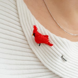May include: A red cardinal bird brooch, crocheted from yarn, is pinned to a white sweater.