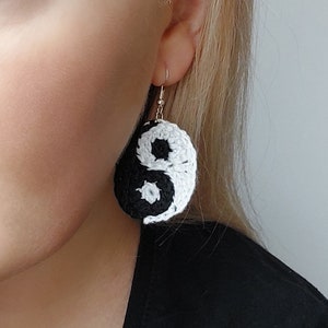 Puede incluir: Pendientes de yin yang de crochet en blanco y negro. Los pendientes cuelgan de la oreja de una persona.
