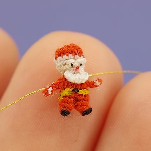 May include: A tiny crocheted Santa Claus figurine in red and white with a yellow belt, holding a gold wire. The figurine is about the size of a fingernail.