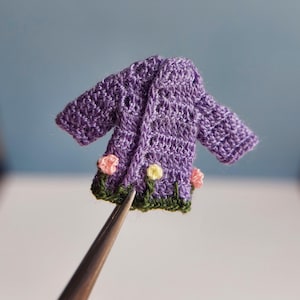 May include: A miniature purple crochet jacket with a floral design. The jacket features a delicate open weave and is adorned with small pink and yellow flowers along the hemline. The jacket is held by a pair of tweezers against a blue background.