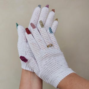 Puede incluir: Un par de guantes de ganchillo blancos con las puntas de los dedos en colores metálicos como plata, rosa, cobre, oro, azul, rojo y morado. Los guantes tienen un puño acanalado y un pequeño anillo dorado en el dedo anular.