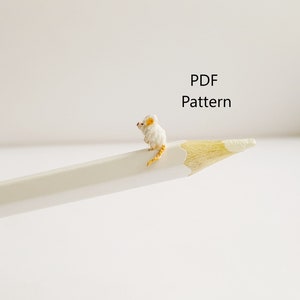 May include: A white crocheted mouse with a yellow tail sitting on a white pencil. The text "PDF Pattern" is written above the mouse.