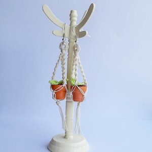 May include: A pair of macrame plant hanger earrings with small terracotta pots and green leaves. The earrings are hanging from a white stand.
