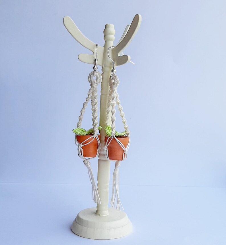 May include: A pair of macrame plant hanger earrings with small terracotta pots and green leaves. The earrings are hanging from a white stand.