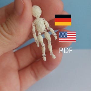 Könnte beinhalten: Eine kleine, cremefarbene Häkelpuppe mit beweglichen Gliedmaßen. Die Puppe wird in einer Hand vor einem hellblauen Hintergrund gehalten. Die Gliedmaßen der Puppe sind mit kleinen, durchsichtigen Perlen verbunden. Das Bild enthält den Text "PDF" und die Flaggen Deutschlands und der Vereinigten Staaten.