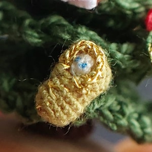 Peut inclure: Un petit berceau crocheté avec une bordure dorée et un bébé bleu et blanc à l'intérieur. Le berceau est niché dans un arbre crocheté vert.