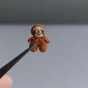 Könnte beinhalten: Eine winzige, handgehäkelte Faultierfigur in Braun- und Beigetönen, mit einem kleinen roten Herz. Das Miniaturfaultier wird von einer Pinzette vor einem neutralen Hintergrund gehalten, was die filigrane Handarbeit zeigt.