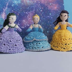May include: Three crocheted dolls in princess dresses. The dolls are in a row, each with a different colored dress: purple, blue, and yellow. The background is a galaxy design. The dolls have different hairstyles and skin tones.