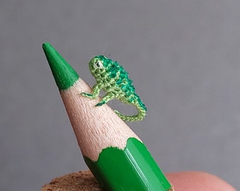 Könnte beinhalten: Ein winziger, handgehäkelter grüner Eidechse, der auf der Spitze eines grünen Bleistifts sitzt. Der Bleistift steht auf einem Korksockel. Der strukturierte Körper und die winzigen Gliedmaßen der Eidechse sind sichtbar. Der Bleistift hat einen Durchmesser von etwa 1,9 cm.