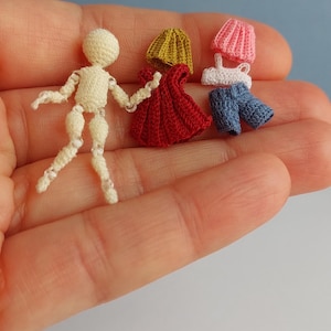 May include: A miniature crochet figure with articulated limbs, next to a red dress, a yellow hat, and a pink hat with a white top and blue shorts. The items are displayed on a person's open palm against a light blue background.