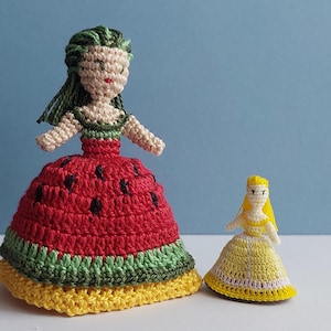 May include: Two handmade crochet dolls. One doll is larger, wearing a red dress with black seed details, green trim, and a yellow base. The other doll is smaller, wearing a yellow dress with white trim. Both dolls have yarn hair and are on a light blue background.