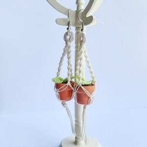 May include: A pair of white macrame plant hanger earrings with small terracotta pots and green leaves. The earrings are hanging from a white stand.