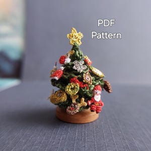 以下が含まれることがあります： 様々なオーナメントが付いた、ミニチュアの編み物クリスマスツリー。金色の星の飾り、赤と金のボール、サンタクロースのフィギュアなど。木製の台座に置かれています。「PDF Pattern」の文字が見えます。