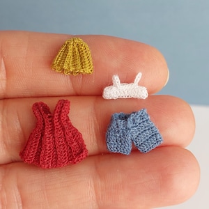 Puede incluir: Conjunto de ropa de ganchillo en miniatura en amarillo, blanco, rojo y azul. El conjunto incluye una falda, un top, un vestido y unos pantalones cortos. Las diminutas prendas están hechas a mano con costuras detalladas, mostrando una artesanía delicada. El fondo es azul claro.