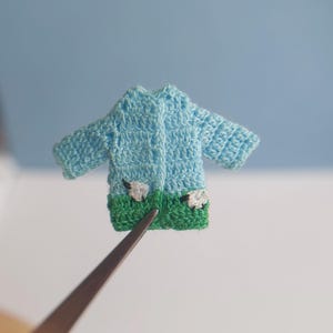 May include: A miniature light blue crochet jacket with green grass and sheep details. The tiny jacket is held by tweezers against a light blue background. The jacket has a button closure.