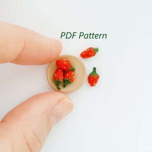 Puede incluir: Un pequeño cuenco de madera lleno de cinco fresas de ganchillo. Las fresas son rojas con hojas verdes. El cuenco está sobre una superficie blanca.