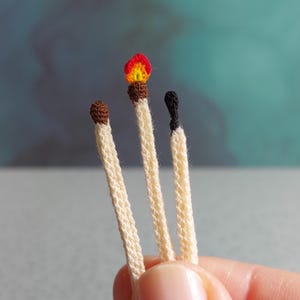 Puede incluir: Primer plano de tres cerillas hechas a mano con ganchillo. Una cerilla está encendida con una llama roja y amarilla, la segunda tiene la cabeza marrón y la tercera la cabeza negra y quemada. Las cerillas son de color blanquecino.