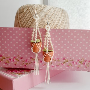 May include: A pair of handmade macrame earrings featuring small terracotta pots and green plants. The earrings are hanging against a pink and white polka dot background.