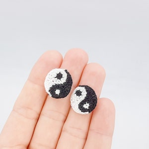 May include: A pair of black and white crocheted yin yang earrings. The earrings are small and round, and they are held in a hand.
